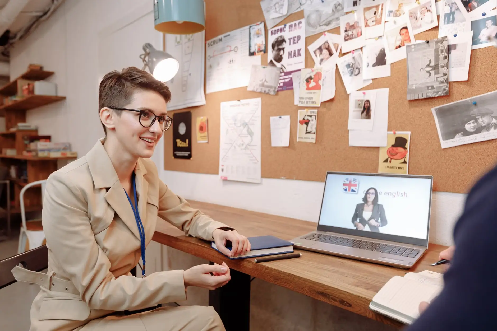 Instrutor sorridente ensina alunos em aula de inglês em sala com quadro de avisos, laptop exibindo “English” e materiais de estudo, destacando Cursos de Inglês: Domine o Idioma (Guia Completo).