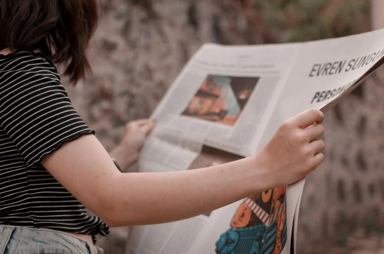 Conteúdos Virais, Dicas e Assuntos do Momento: Mulher lendo um jornal ao ar livre, segurando a página com as mãos, com foco nas letras e ilustrações da edição em destaque.