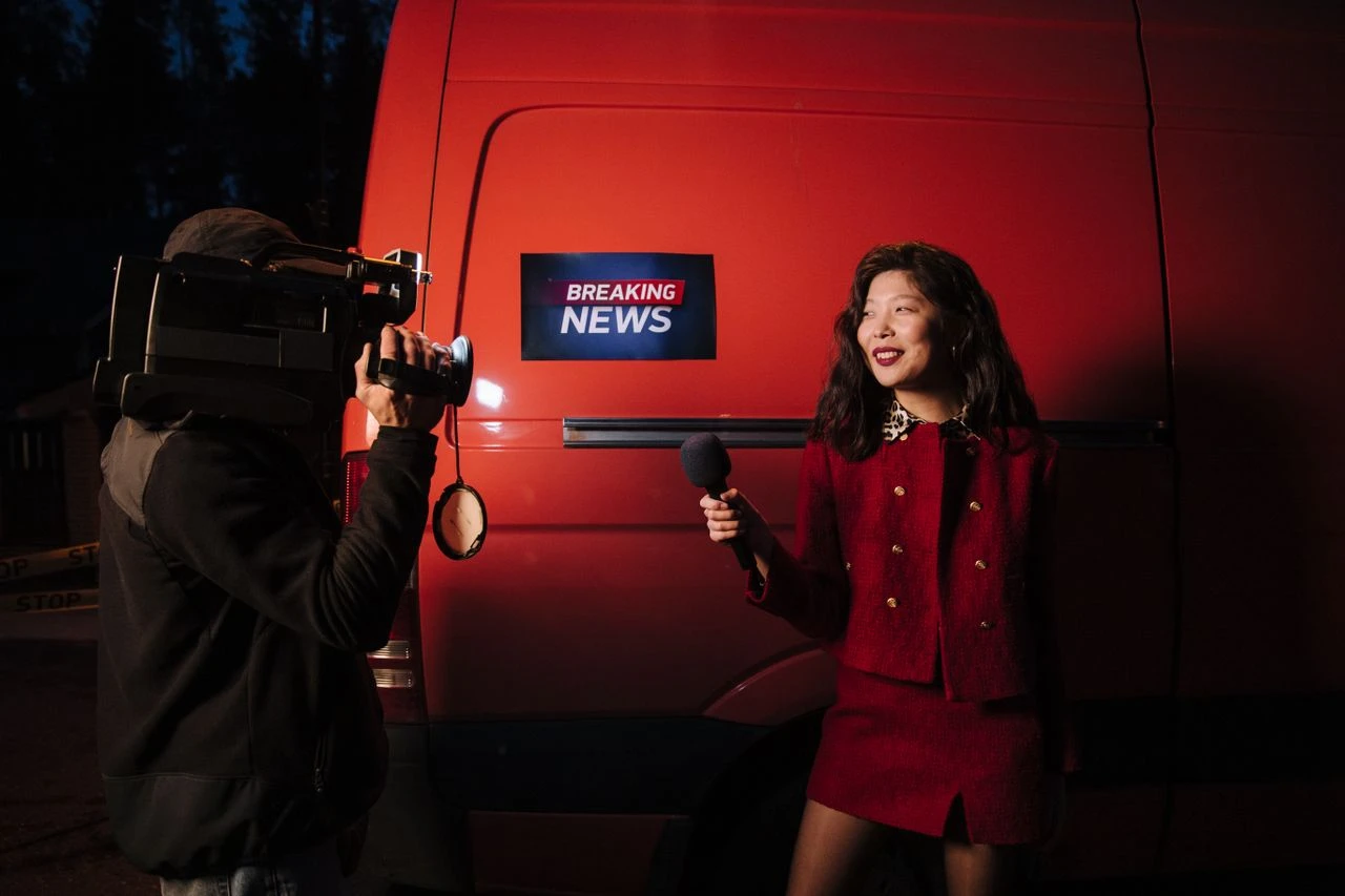 Entretenimento e informação: Conteúdos que mantêm você sempre atualizado: Câmera de reportagem em primeiro plano enquanto uma entrevistada, em trajo vermelho, aparece ao lado de um veículo com a placa de “BREAKING NEWS”.