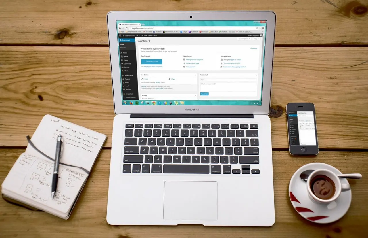 Qual o melhor site gratuito para criar um Blog? Confira as melhores opções: Notebook com tela exibindo painel de trabalho, celular ao lado e xícara de café sobre mesa de madeira, ambiente de produtividade para criar um blog